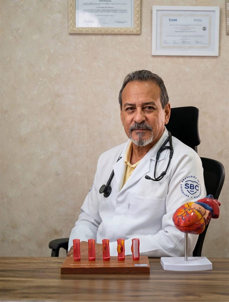 Dr. Abraão Telles Rocha, cardiologista, sentado com jaleco branco e estetoscópio, segurando um modelo anatômico do coração ao lado de modelos de stent arterial
